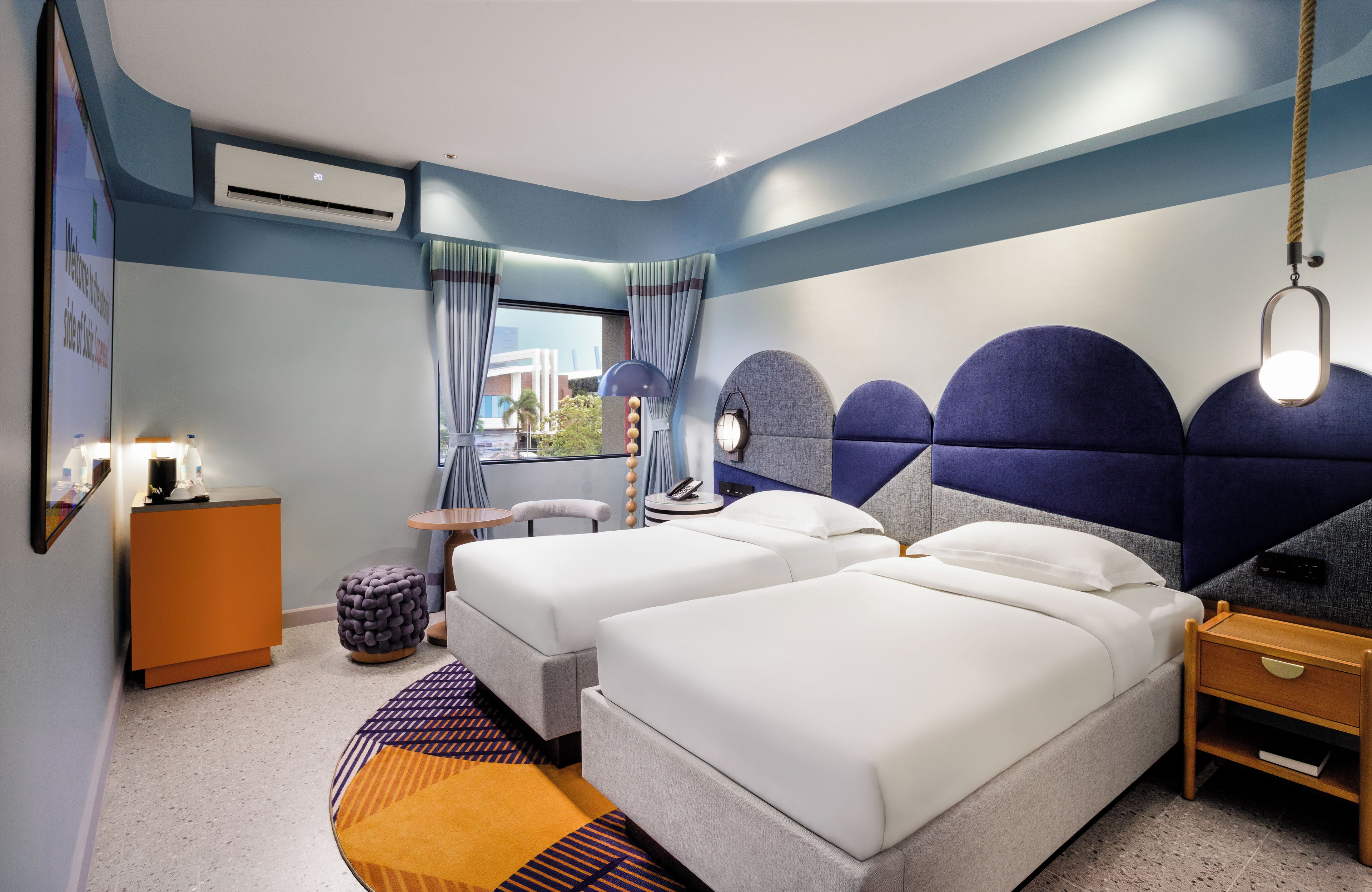 A modern hotel room with two beds, a desk, a chair, a small ottoman, and a nightstand.