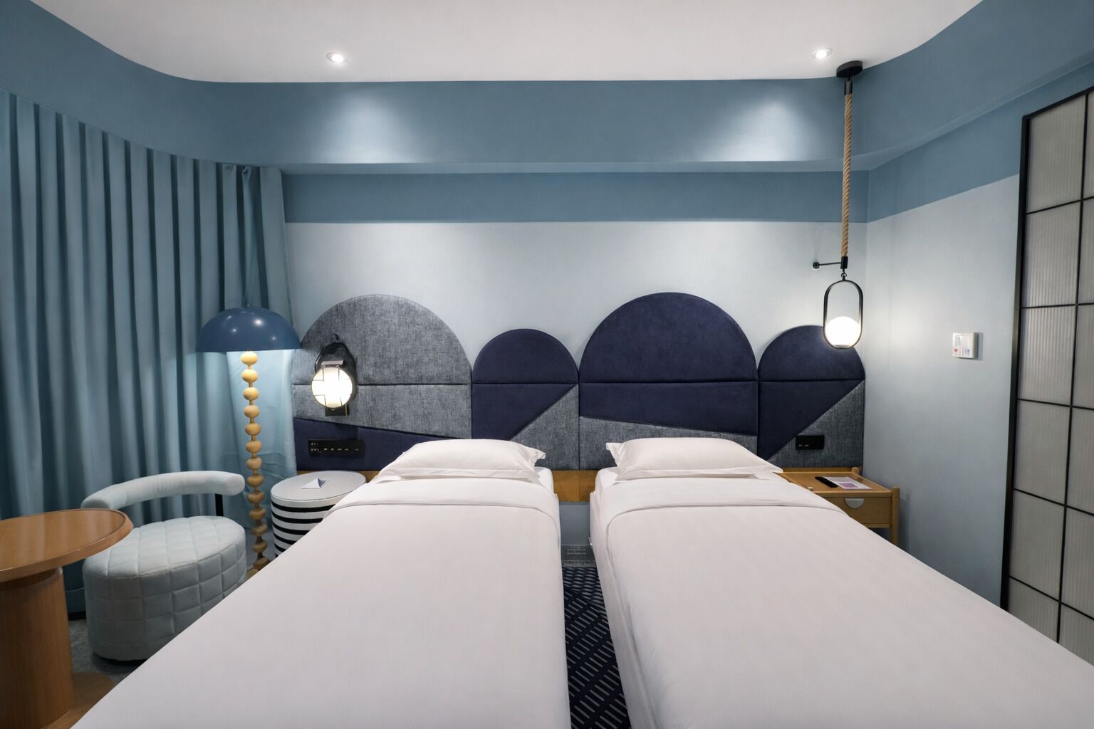 A hotel room with two single beds, a desk, a chair, a lamp, and a large window.