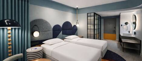 Habitación de hotel con dos camas, un escritorio, una silla, una mesa redonda y un baño con espejo y lavamanos.