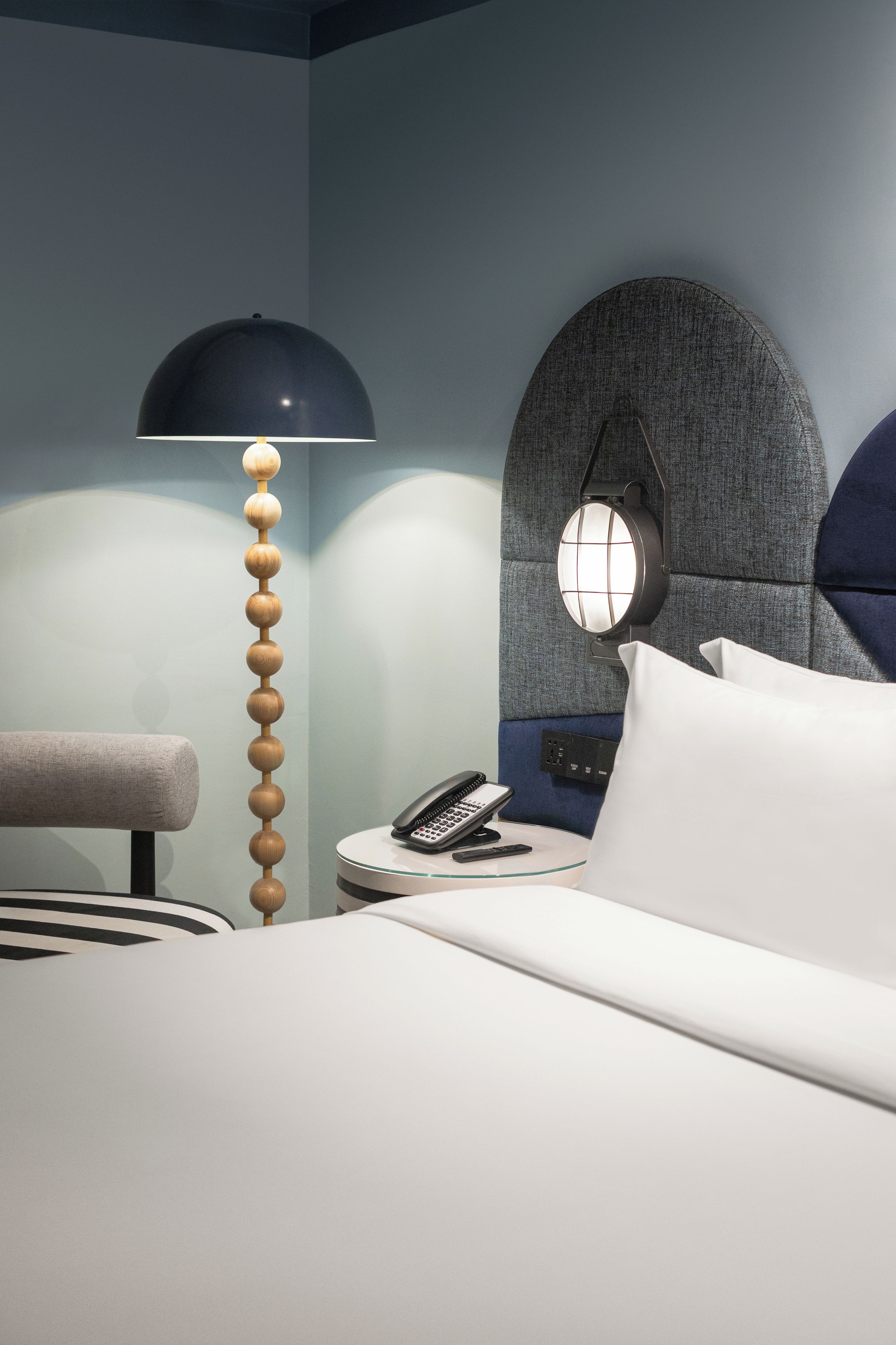 A modern hotel room with a bed, a desk with a telephone, a lamp, and a wall-mounted light fixture.