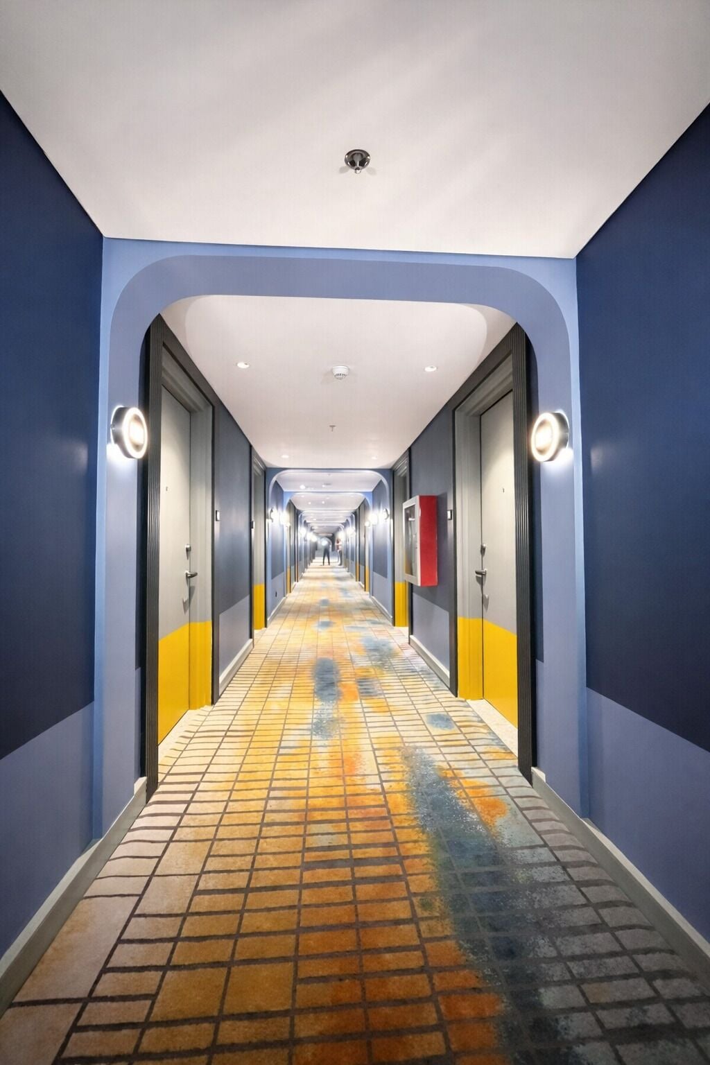 A hotel corridor with a tiled floor, blue walls, and doors with light fixtures.