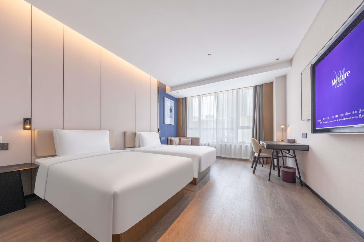 A modern hotel room with two beds, a desk, and a large screen displaying the hotel name.