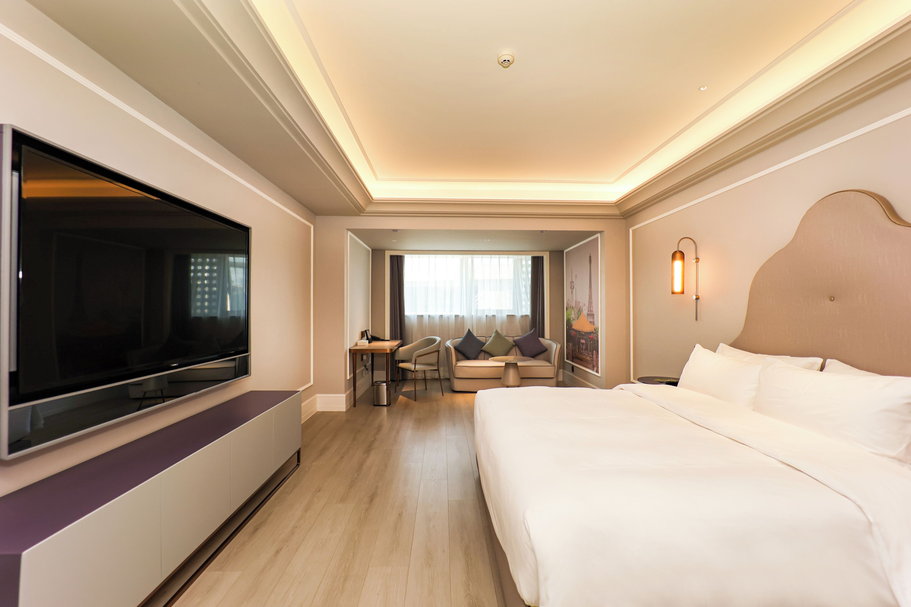 Une chambre d’hôtel dotée d’un grand lit, d’un téléviseur à écran plat fixé au mur, d’un coin salon avec une table et des chaises, et d’une fenêtre avec des rideaux.