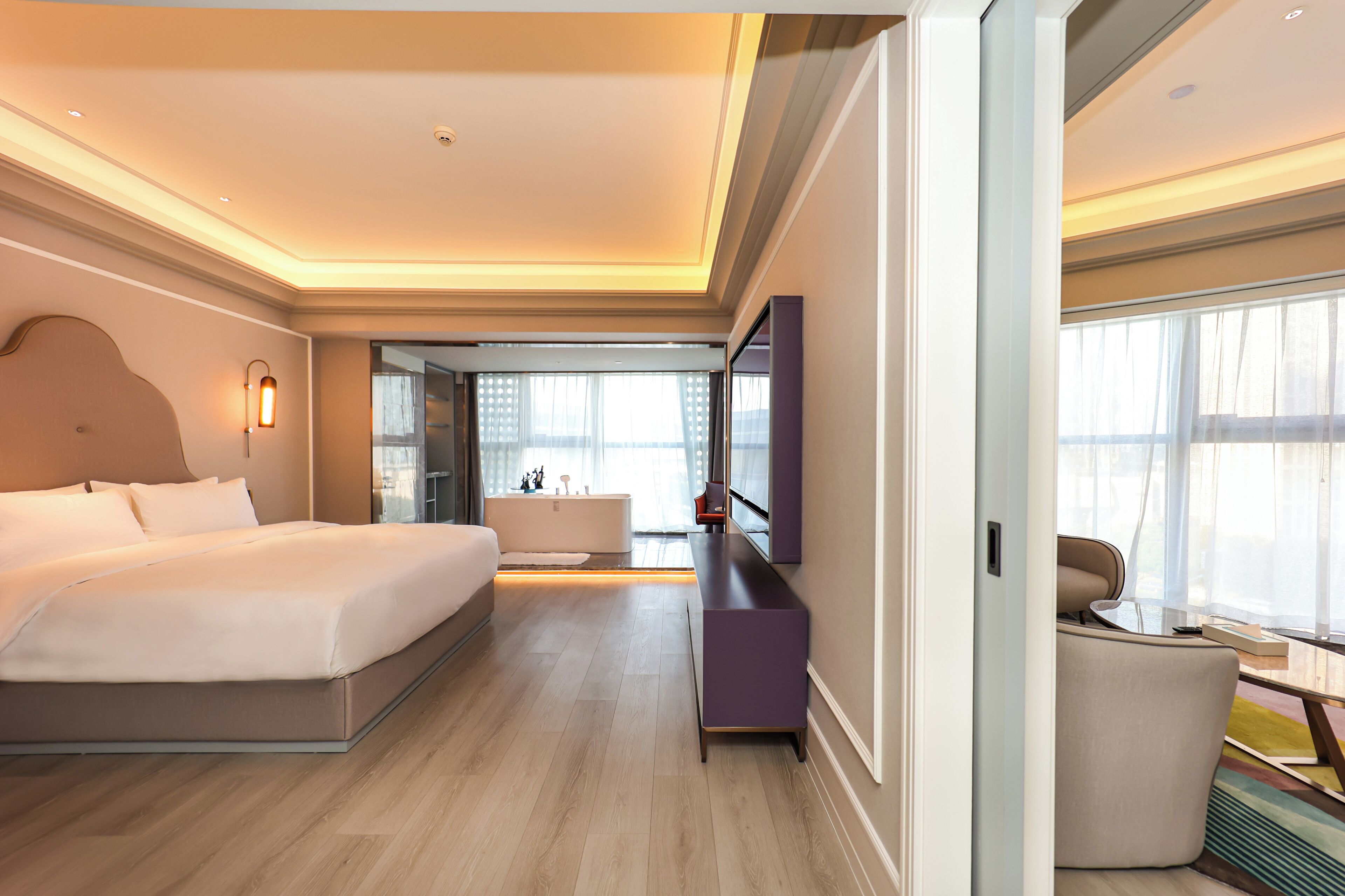 A modern hotel room with a large bed, a sitting area with a chair, a small table, a bathtub, and a large window.