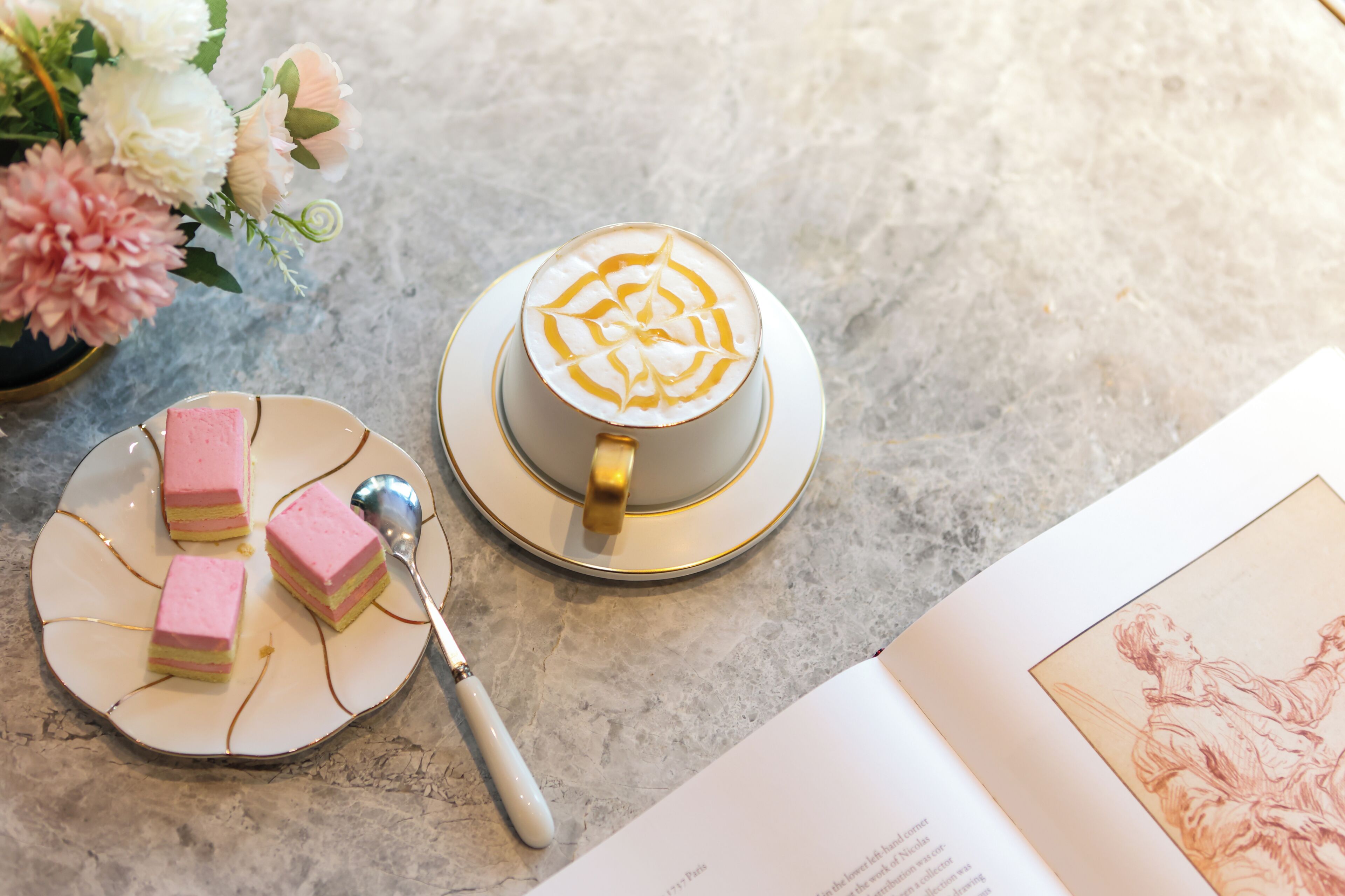 Une tasse de café ornée d’un motif en latte art, une assiette avec des cubes roses, une cuillère et un livre illustré, le tout posé sur une table.