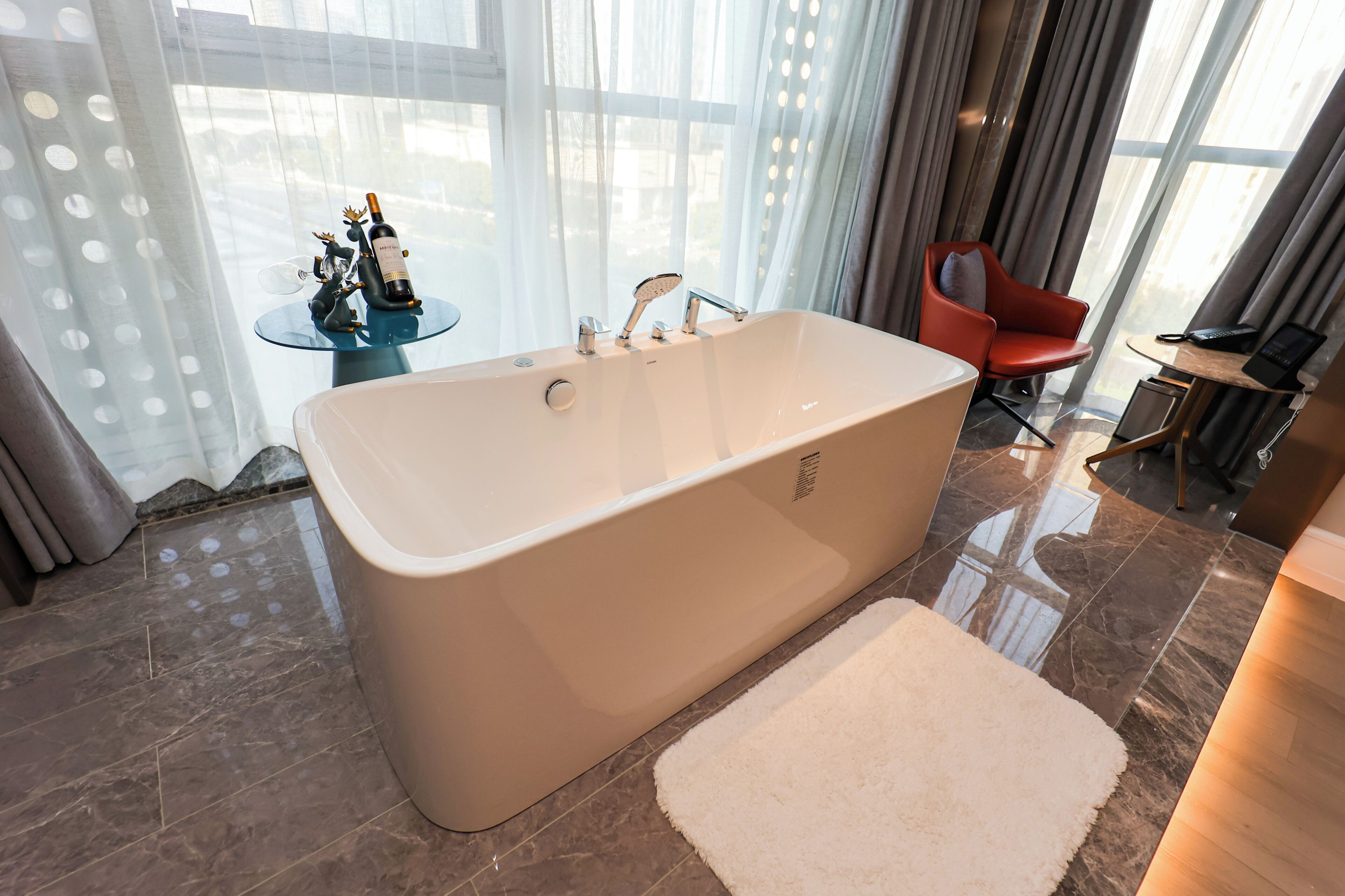 A modern bathroom with a freestanding bathtub, a small round table with a bottle and figurines, a red chair, and a desk with a phone.