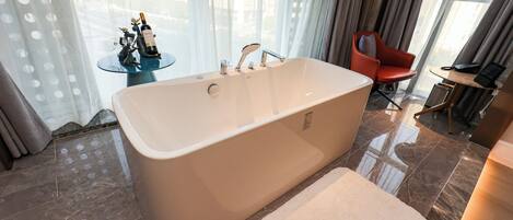 A modern bathroom with a freestanding bathtub, a small round table with a bottle and figurines, a red chair, and a desk with a phone.