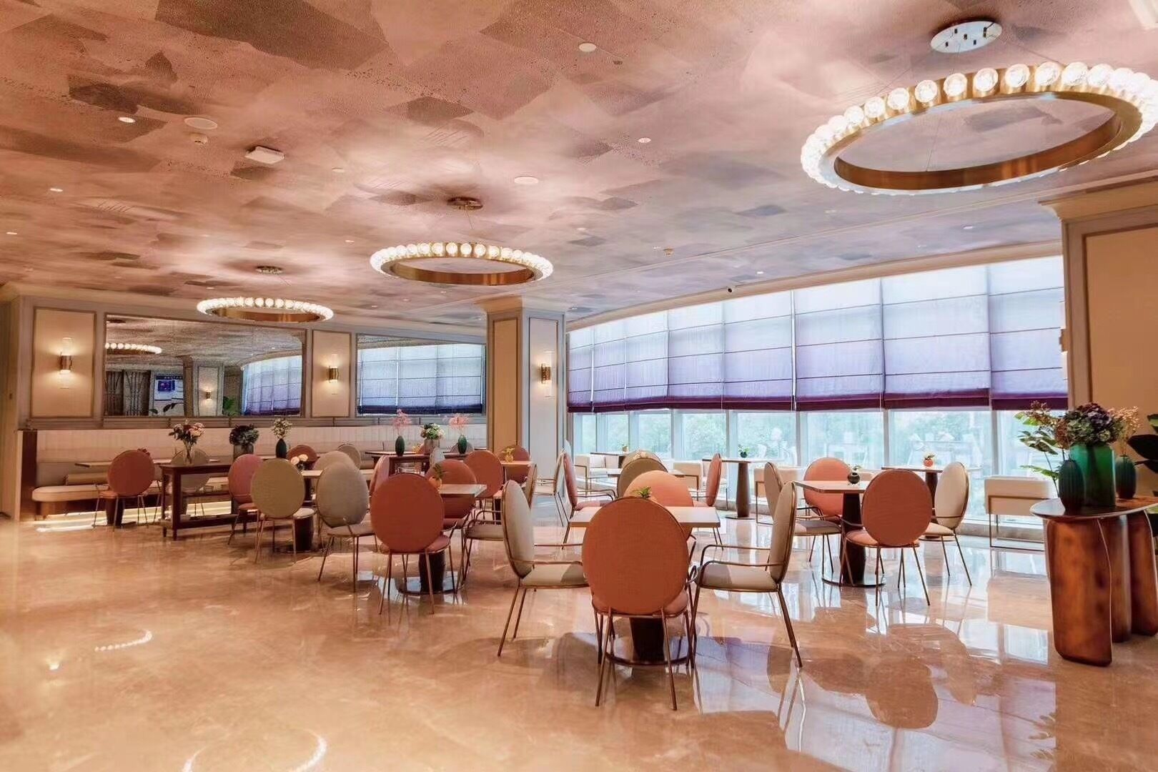 A spacious hotel lobby with multiple dining tables and chairs, large windows, and a modern ceiling design.