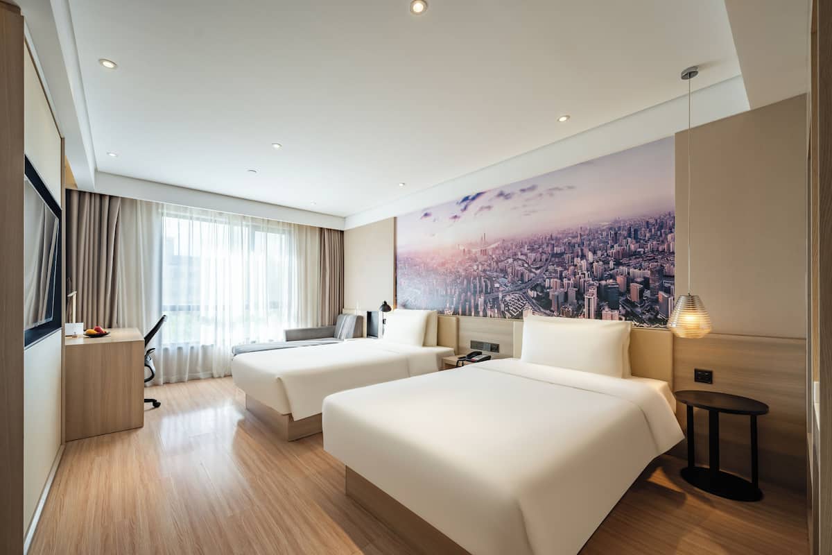 A hotel room with two beds, a desk, a chair, a lamp, and a cityscape mural on the wall.