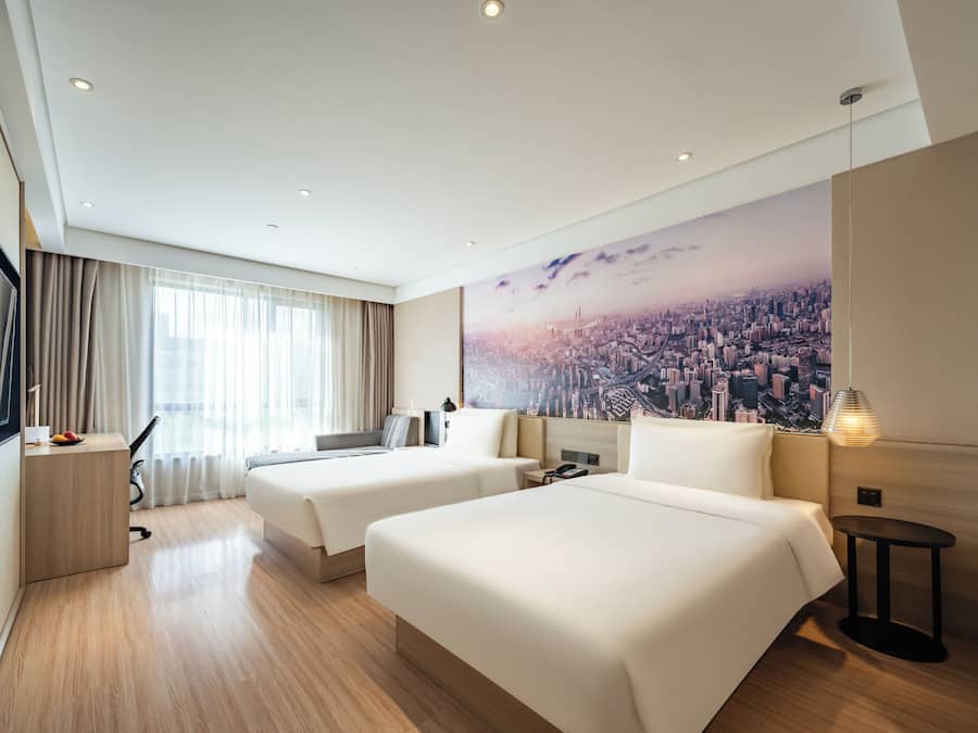 A hotel room with two beds, a desk, a chair, a lamp, and a cityscape mural on the wall.