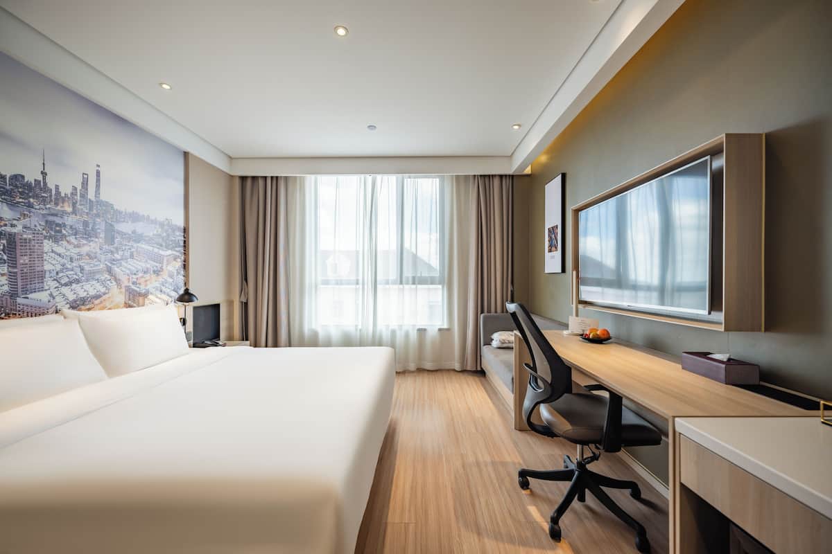 A hotel room with a large bed, a desk with a chair, a television, and a cityscape mural on the wall.