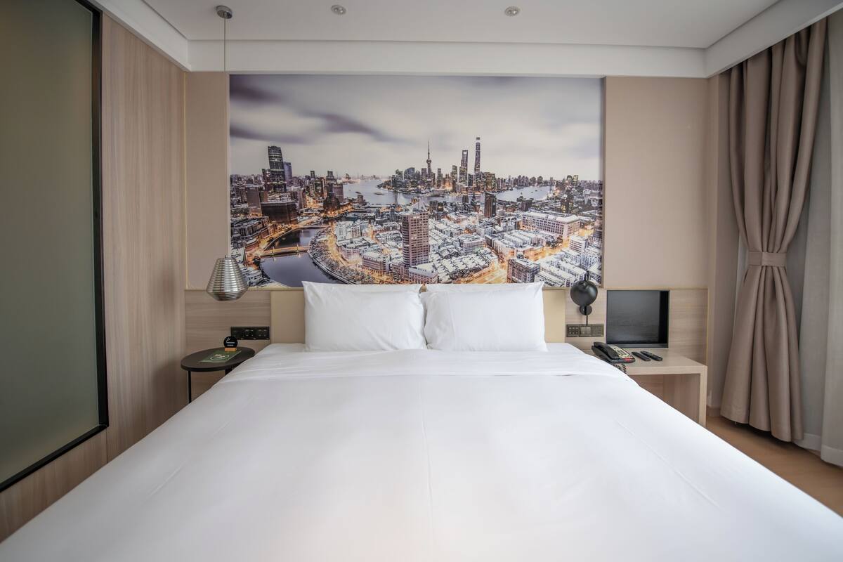 A hotel room with a large bed, a nightstand with a lamp, a TV, and a cityscape mural on the wall.