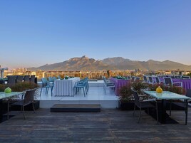 A rooftop terrace with tables and chairs, a view of a mountain, and a cityscape in the background.