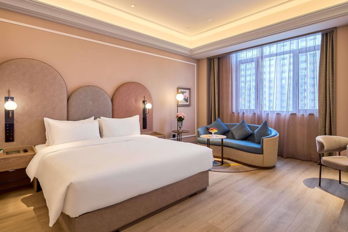 Une chambre d’hôtel avec un grand lit, un canapé, un fauteuil, une petite table et une grande fenêtre avec des rideaux.