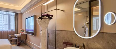 A hotel room with a bed, a desk, a large mirror, a bathroom with a sink, and a shower area.