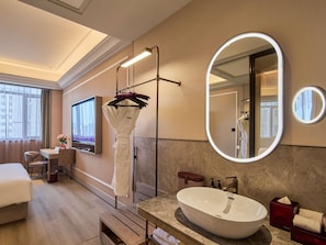 Habitación de hotel con cama, escritorio, espejo grande, baño con lavamanos y zona de ducha.
