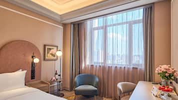 A hotel room with a bed, two chairs, a small table with a vase of flowers, a desk with a sign, and a window with curtains.