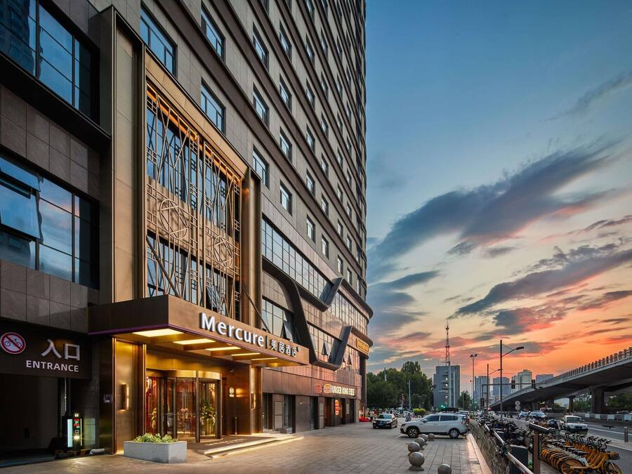 Um hotel moderno com entrada destacada, placa visível do 'Mercure' e paisagem urbana ao fundo.