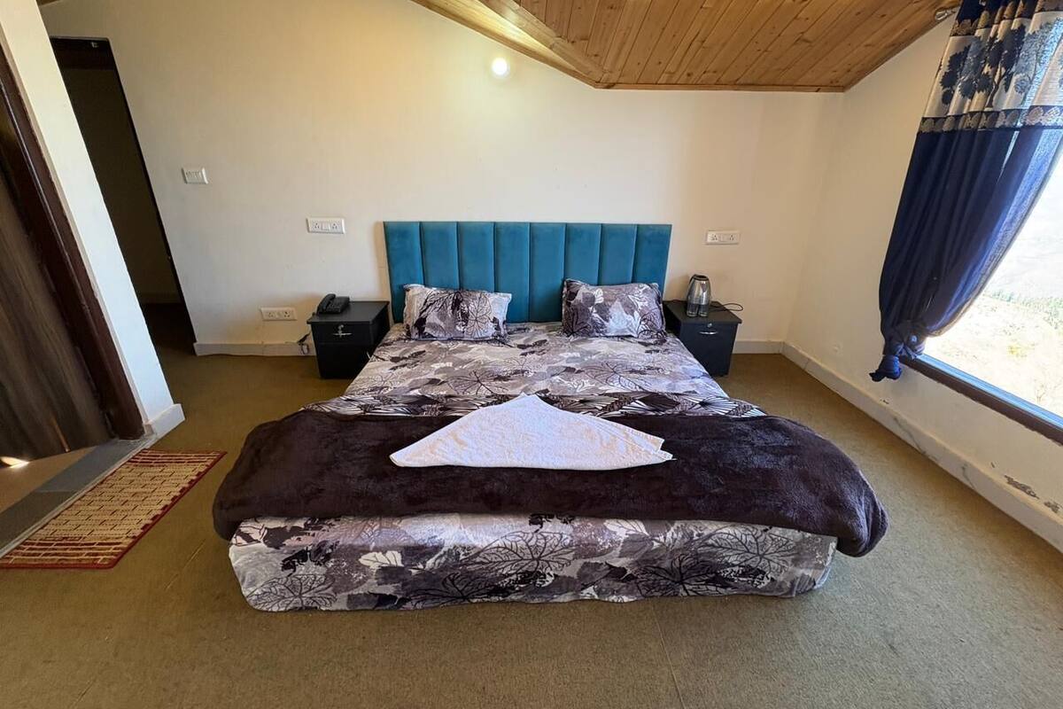 A large bed with a blue headboard, a patterned bedspread, and a dark throw blanket. There is a bedside table with a lamp and a small decorative item. The room has a wooden ceiling and a window with curtains.