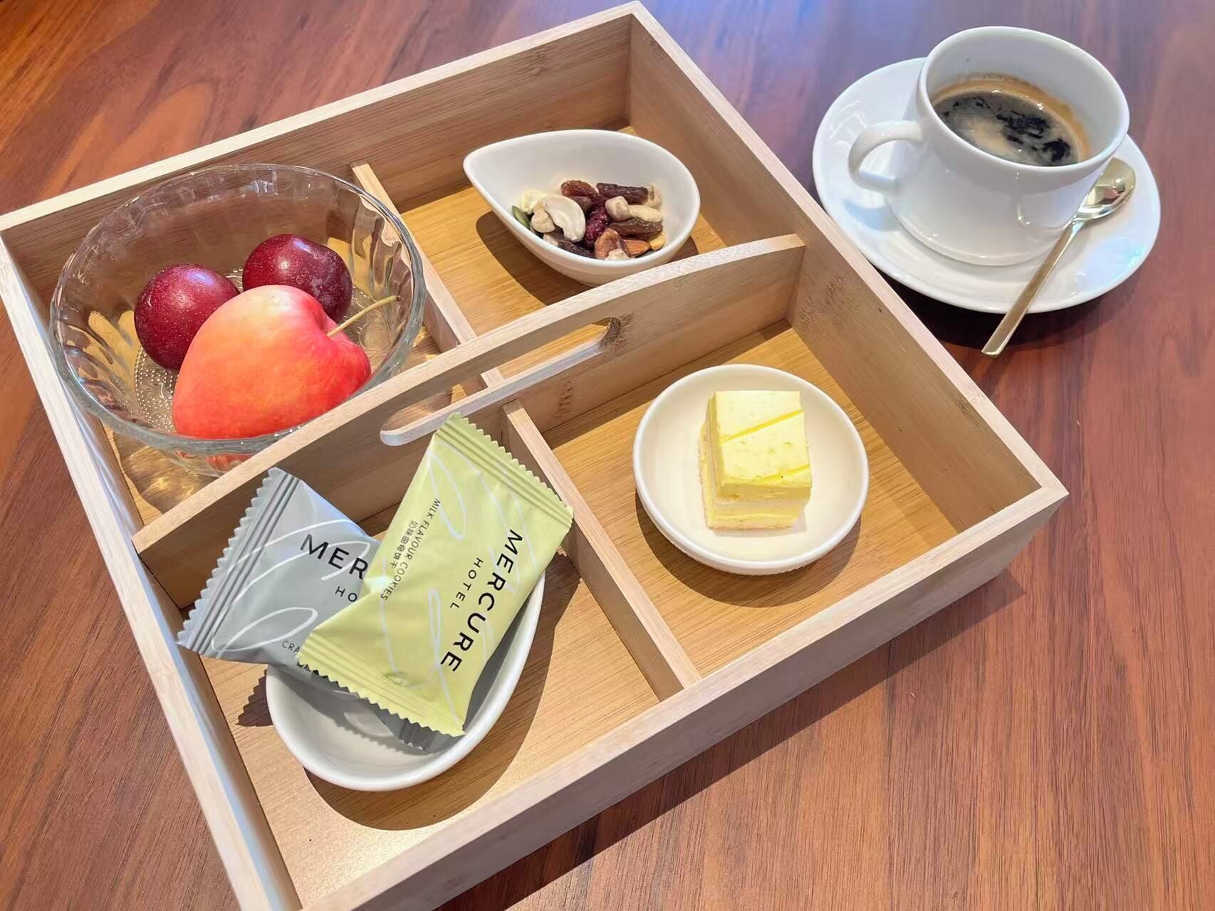 A wooden tray with a glass bowl of fruit, a bowl of nuts, a packet of tea, a slice of butter, and a cup of coffee.
