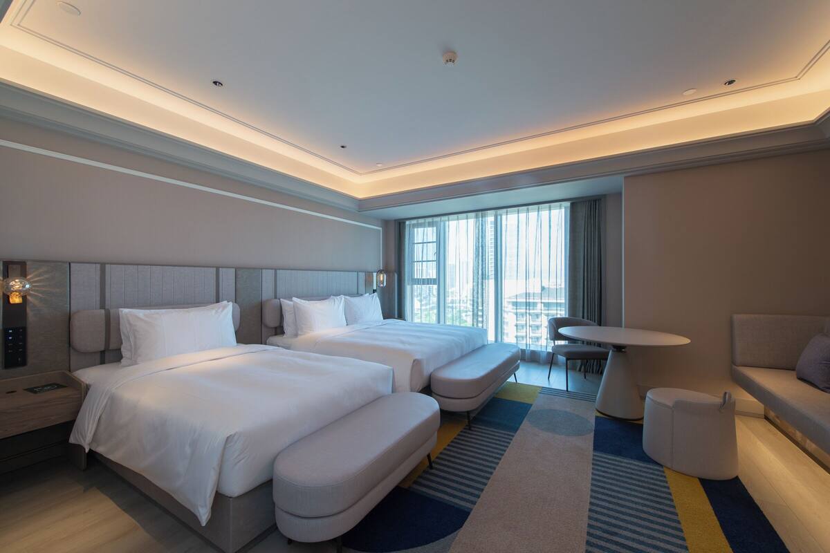 Una habitación de hotel moderna con dos camas, un sofá, una mesita y vistas a la ciudad a través de amplios ventanales.
