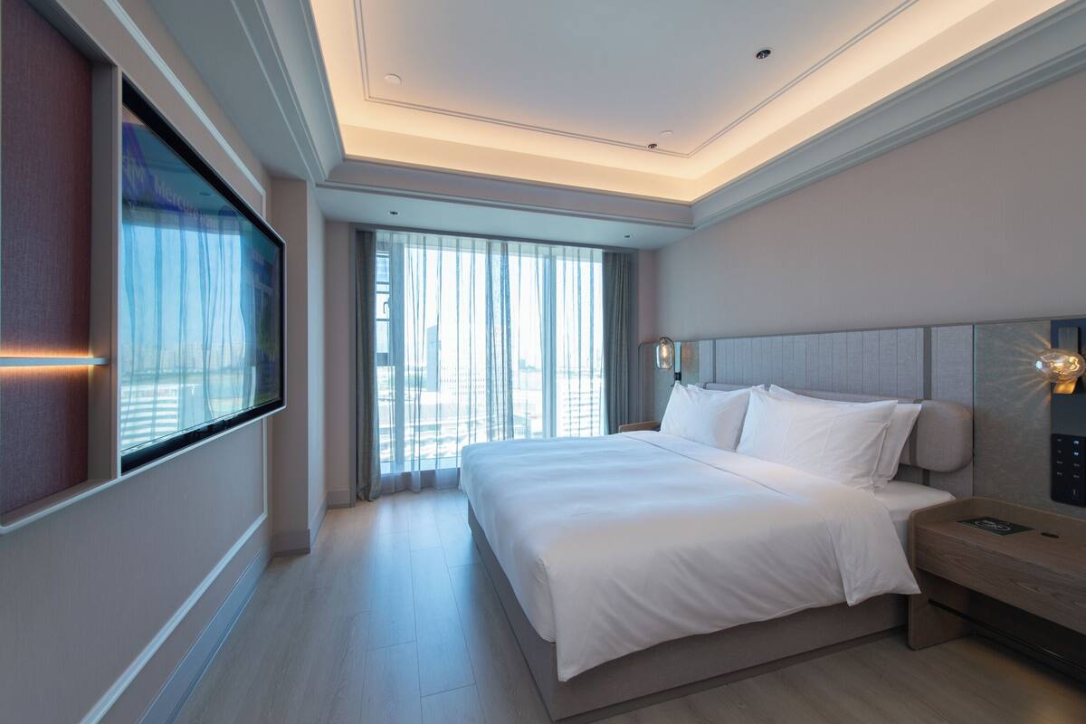 Una habitación de hotel moderna con una cama grande, un televisor montado en la pared y vistas de la ciudad a través de amplias ventanas.