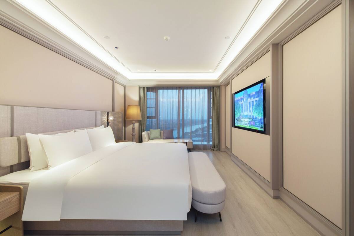 Una habitación de hotel moderna con una cama grande, una mesita de noche, un televisor y vistas a la ciudad desde la ventana.