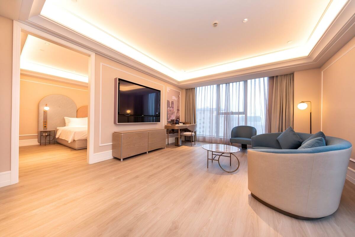 Een moderne hotelkamer met een flatscreen televisie, een bank, een eettafel en een groot raam met gordijnen.