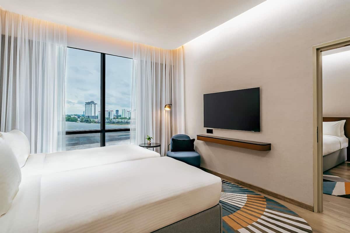 A hotel room with a bed, a TV mounted on the wall, a small table, and a view of the cityscape through large windows.