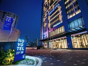A hotel entrance with illuminated signage, a paved walkway, and a cityscape in the background.