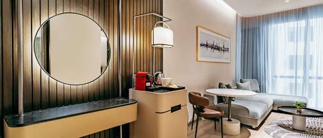 Una habitación de hotel moderna con pared de paneles de madera, un espejo redondo, un escritorio con una cafetera roja, un sofá, una mesa de comedor y un ventanal con cortinas.