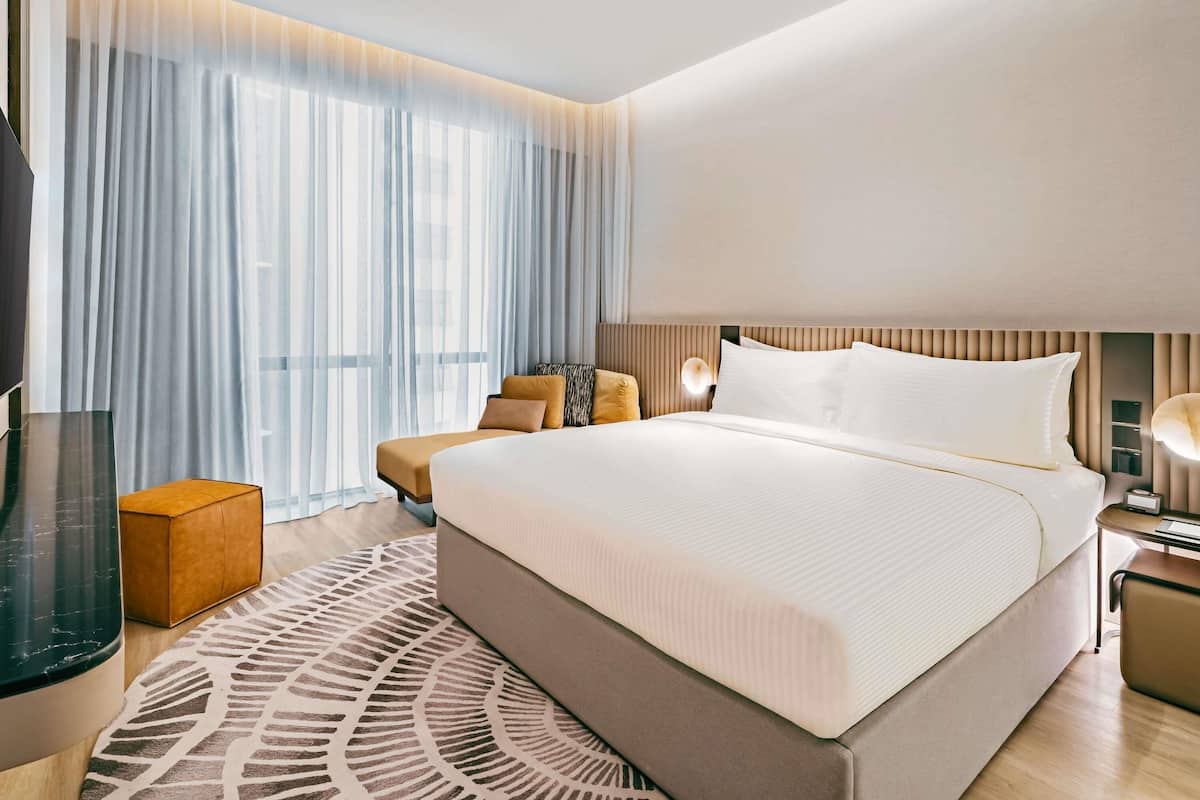 A modern hotel room with a large bed, a bedside table, a lamp, and a view of the city through sheer curtains.