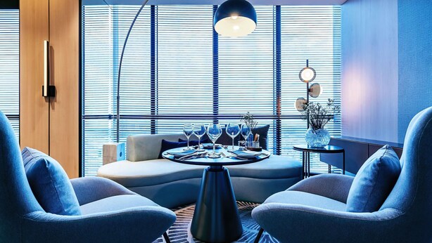 Una habitación moderna con sofá, dos sillones, una mesa de comedor con vasos y un amplio ventanal con persianas.