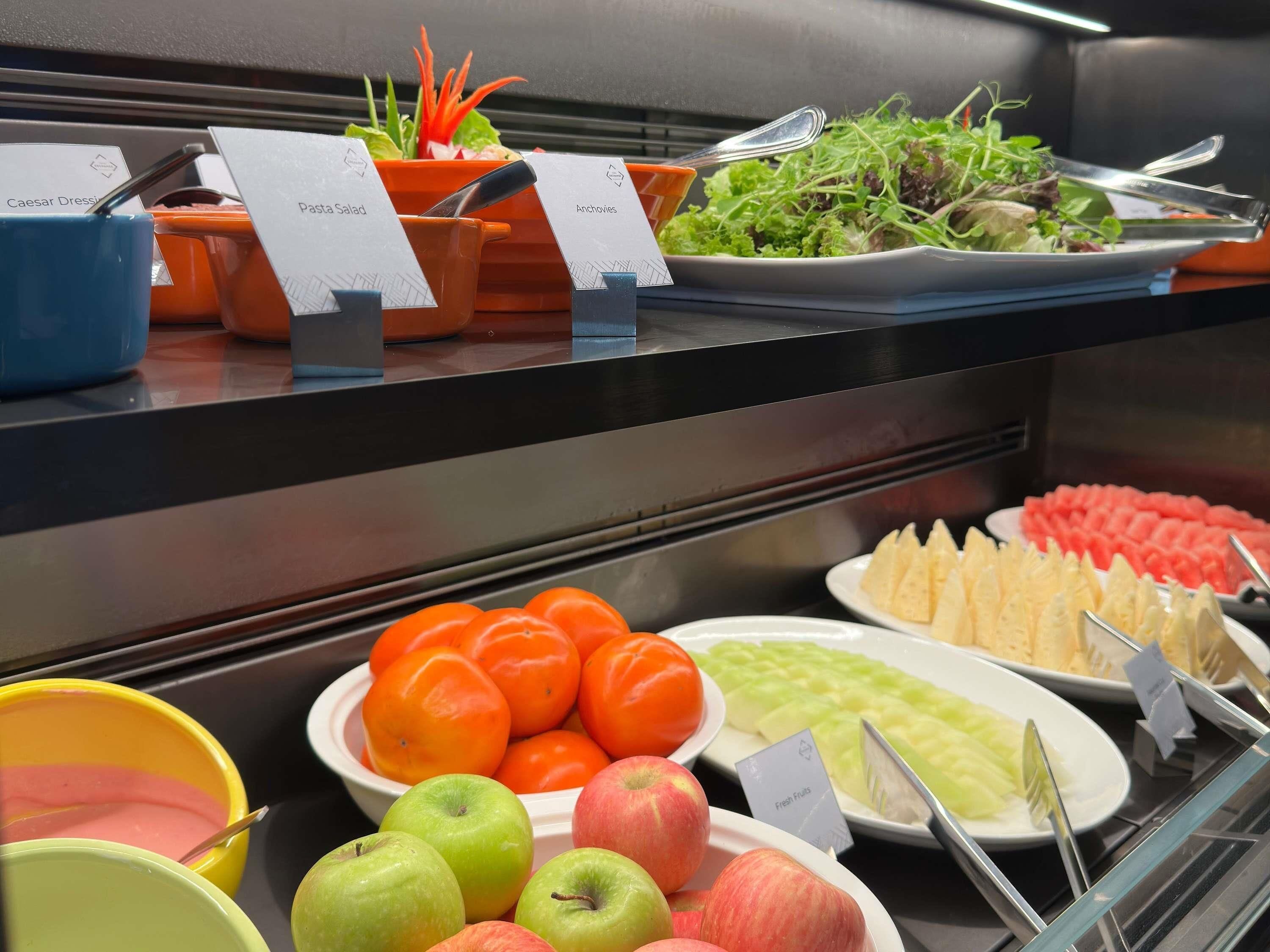 Buffet com pratos identificados, incluindo molho Caesar, salada de macarrão, anchovas e frutas frescas.