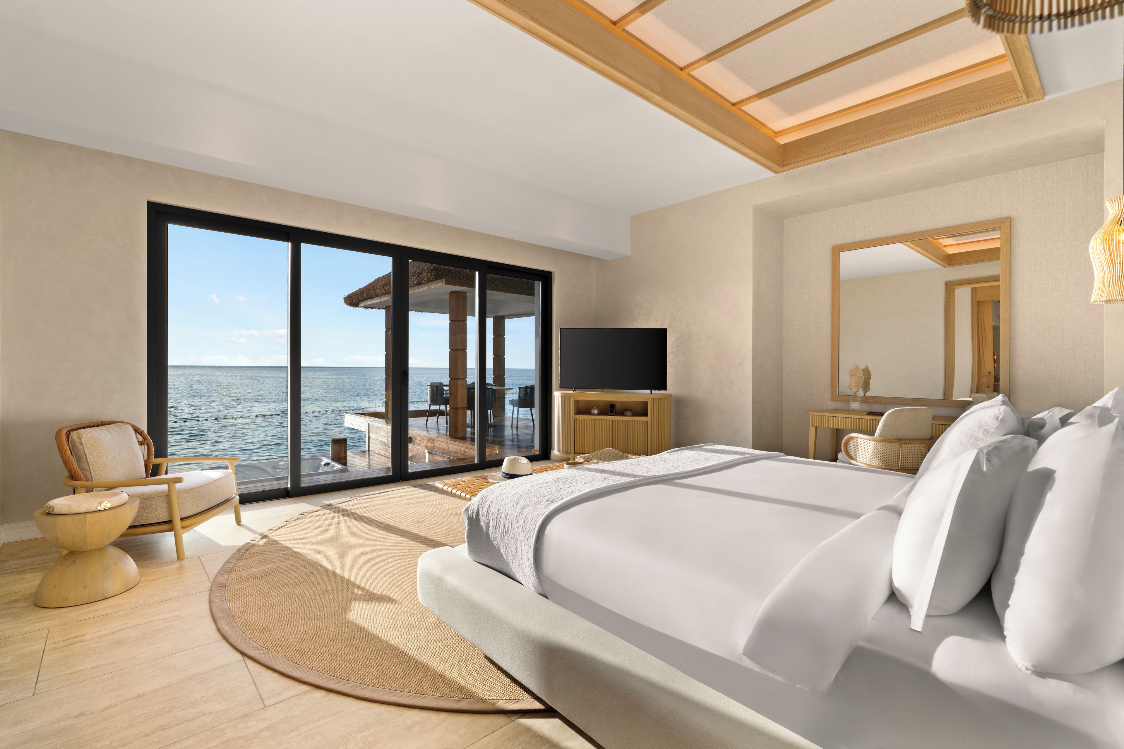 A bedroom with a large bed, a television, a mirror, and a view of the sea through a sliding glass door.