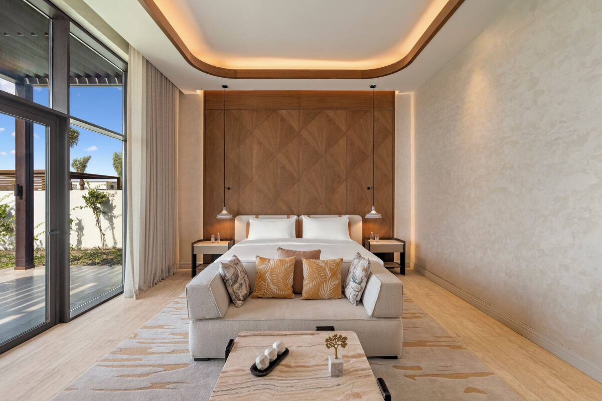 Un dormitorio moderno con una cama grande, un sofá y una mesita de café. Hay dos mesitas de noche y un ventanal con vistas a palmeras.