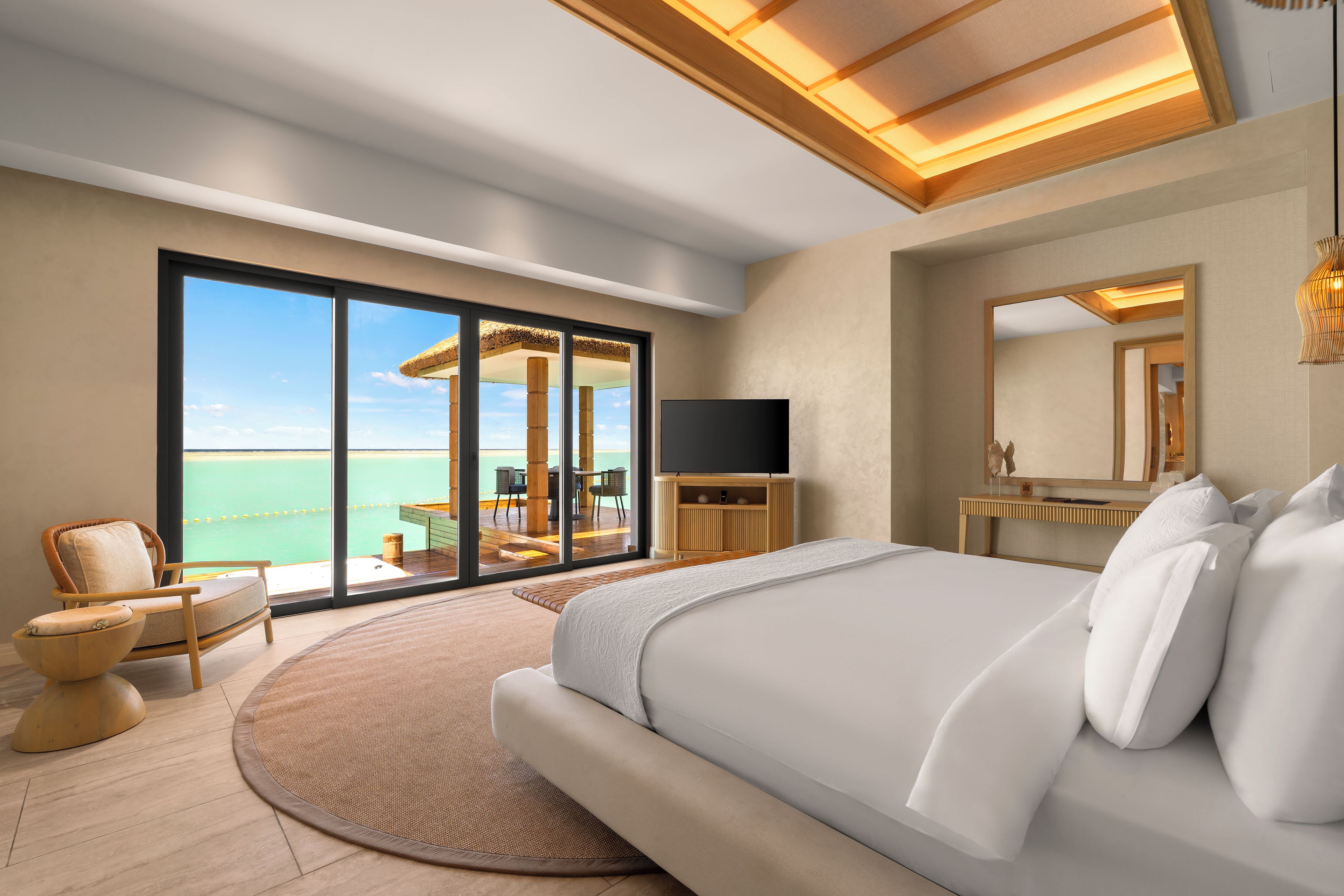 A modern bedroom with a large bed, a TV, a desk, and a view of the ocean.