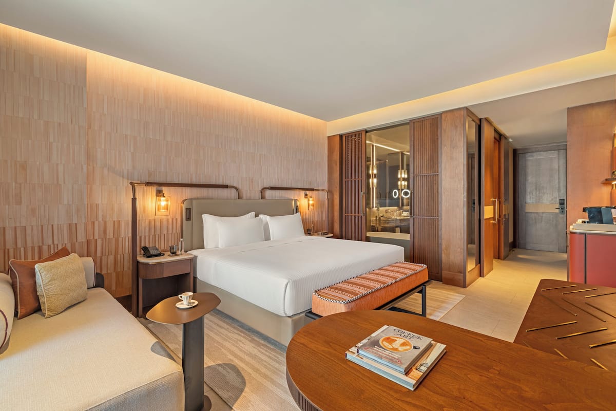 Una habitación de hotel moderna con una cama grande, mesitas de noche, una zona de estar y vistas a la cocina.