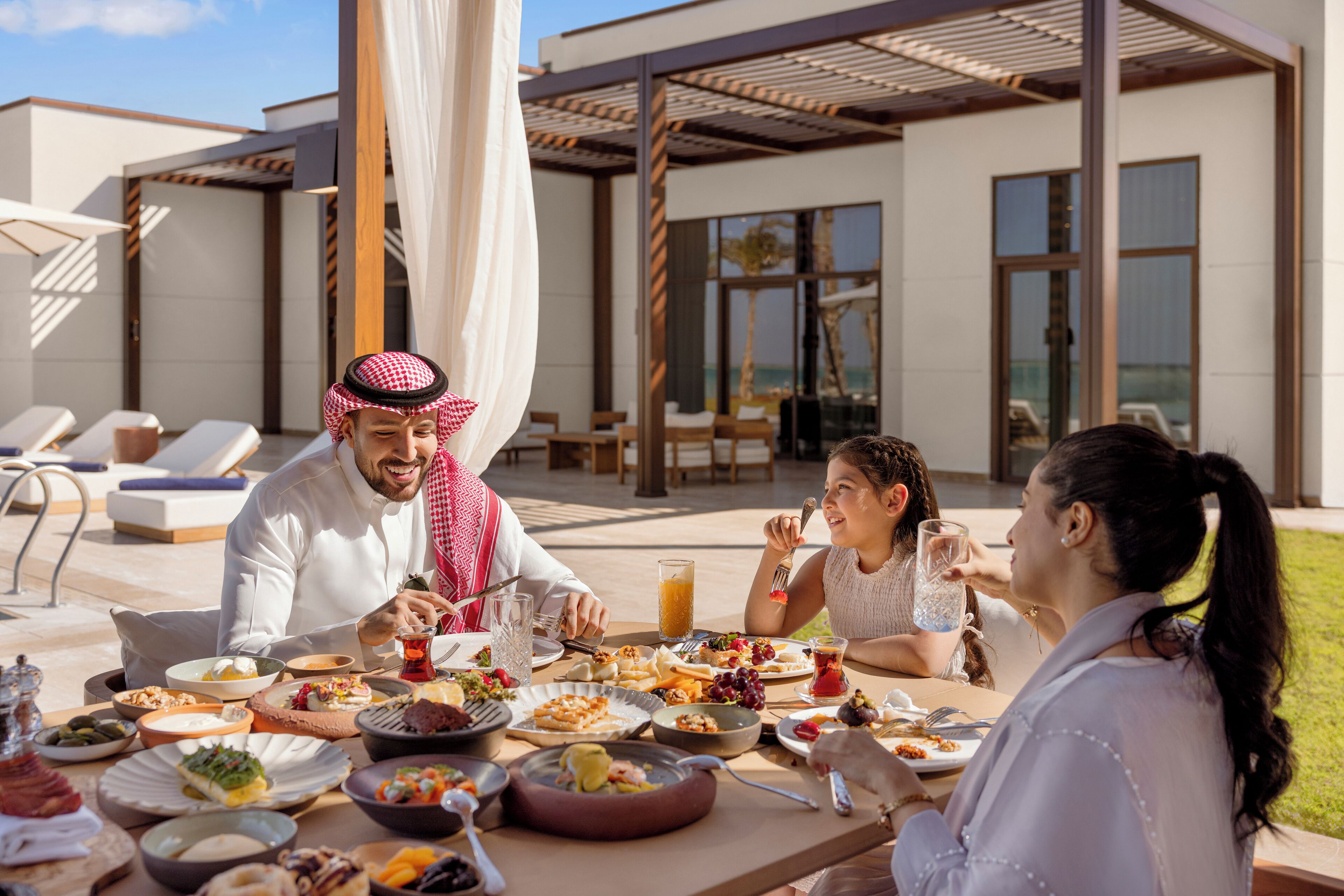 Una mesa de comedor con diversos platos y bebidas, ubicada al aire libre con un edificio moderno al fondo.