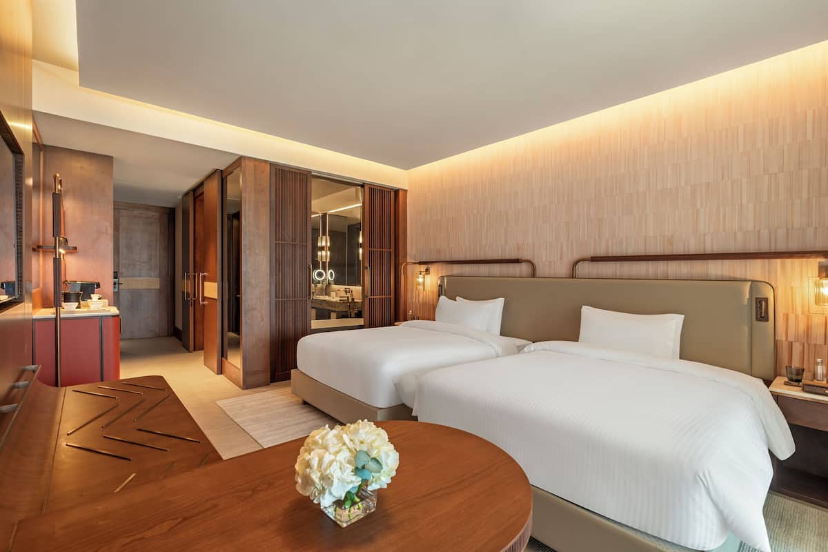 Habitación de hotel con dos camas, un escritorio de madera, un espejo y un baño visible a través de una puerta abierta.