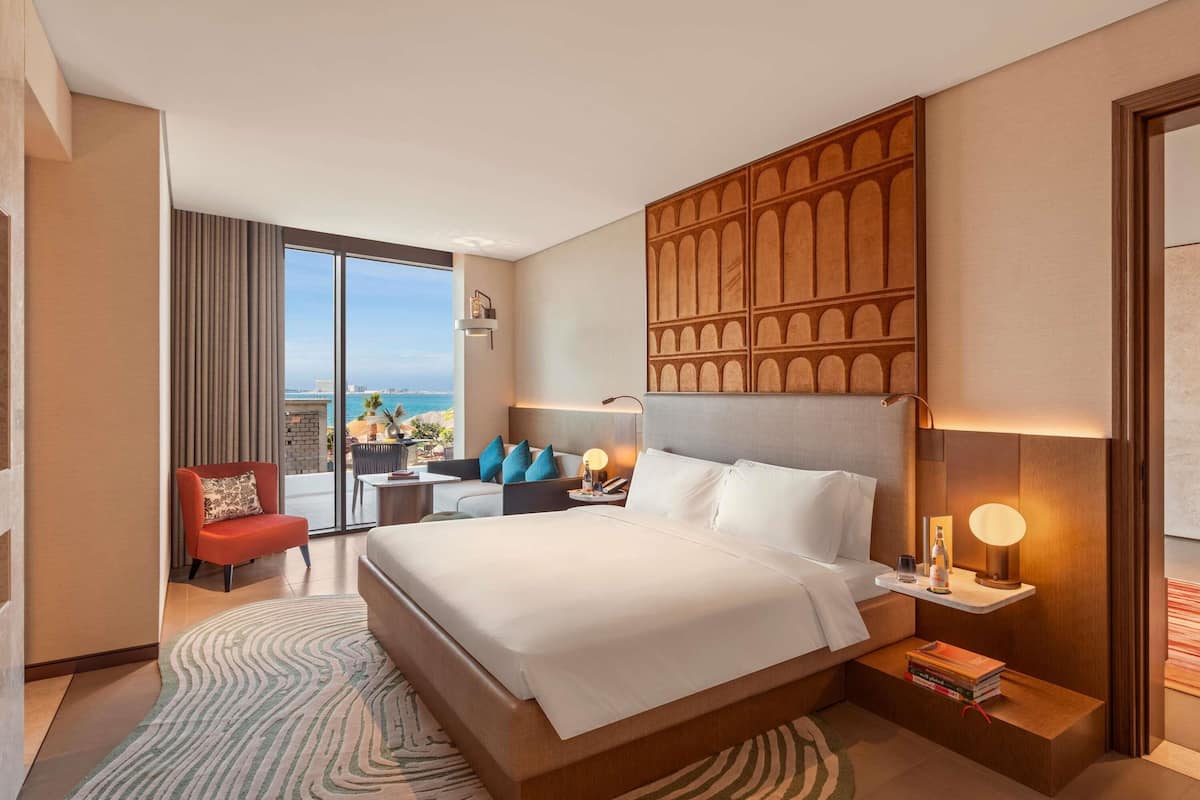 A hotel room with a large bed, a seating area with a view, a small table with books, and a nightstand with a lamp.