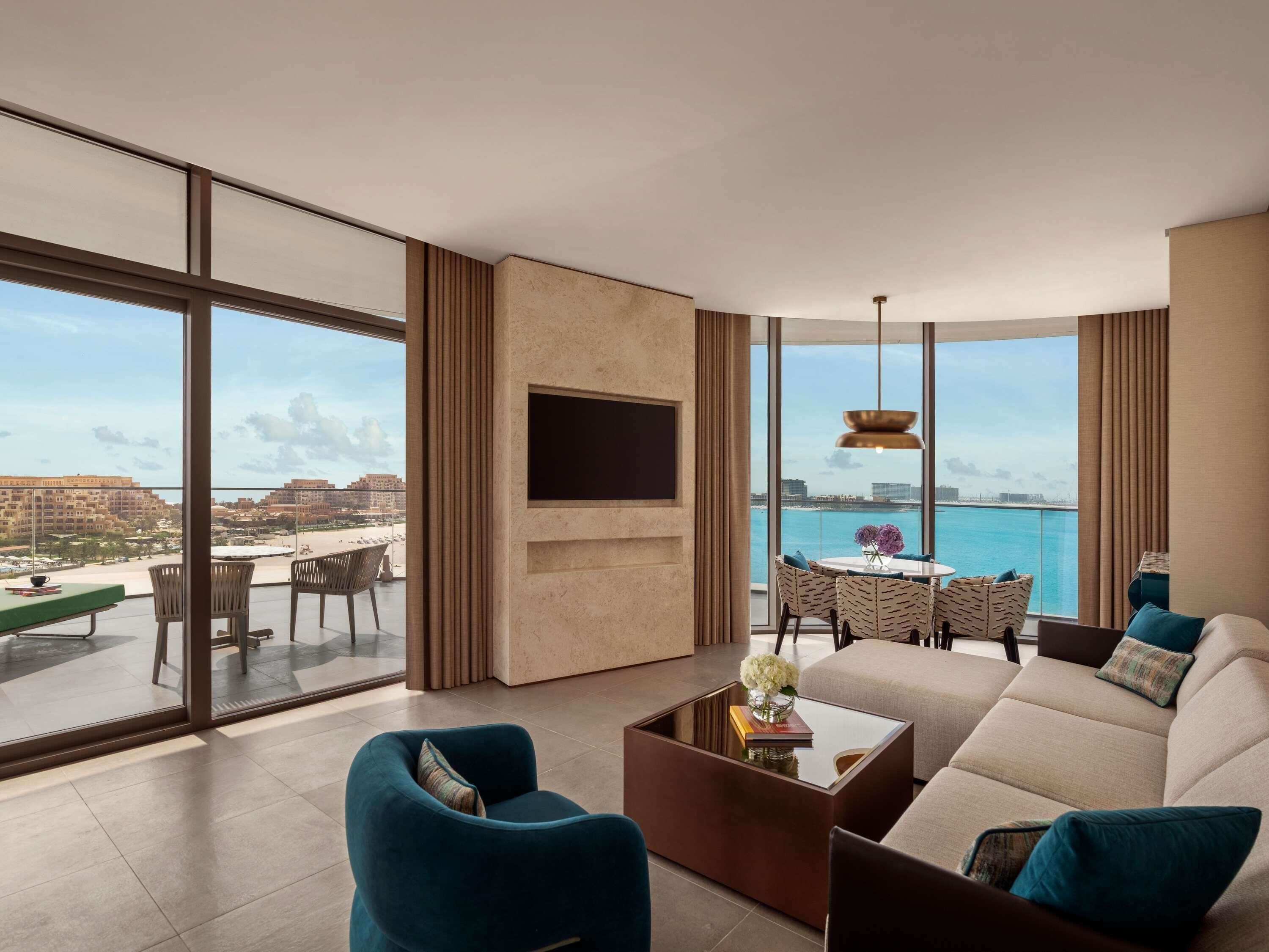A modern living room with a large TV, comfortable seating, a dining area, and a view of the sea.