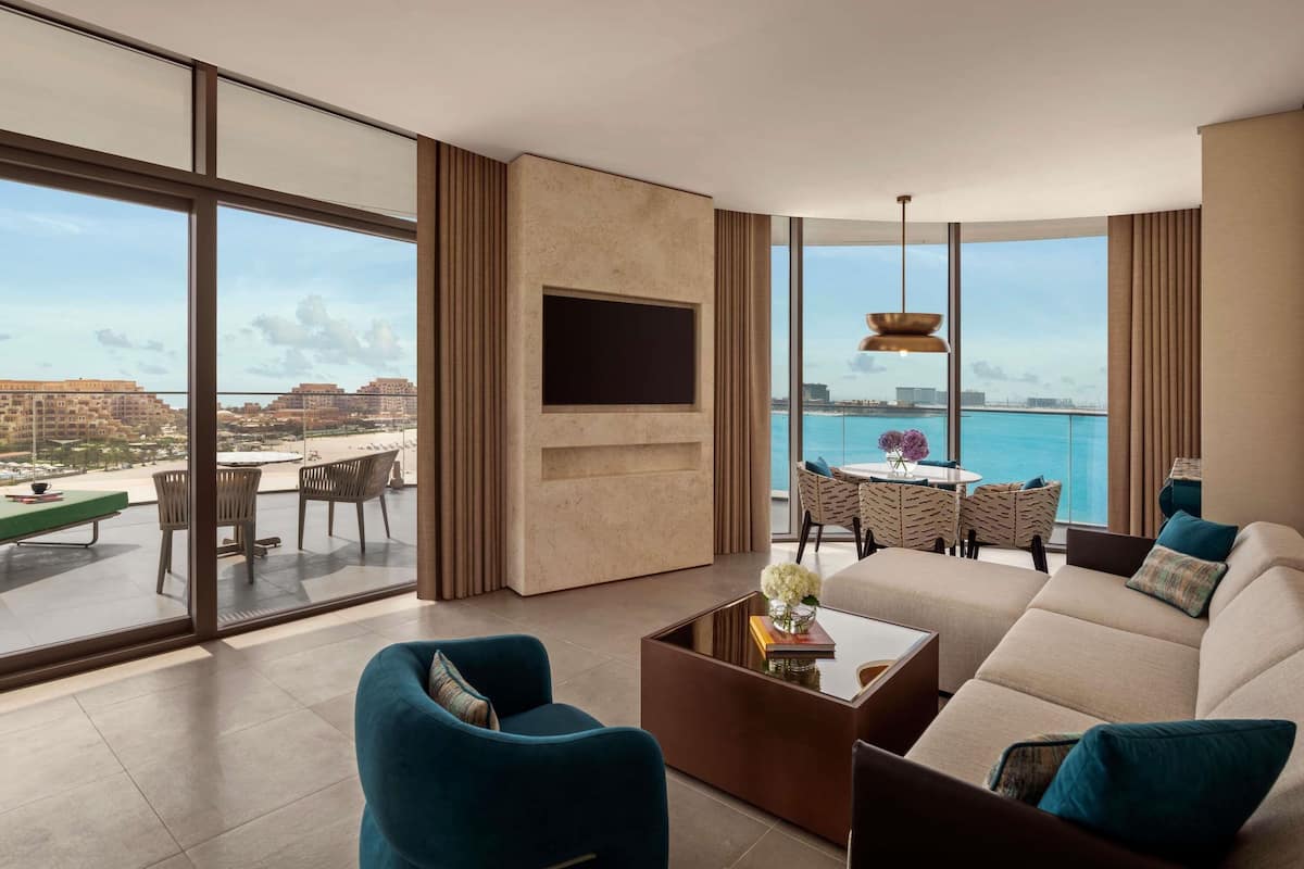 A modern living room with a large TV, comfortable seating, a dining area, and a view of the sea.