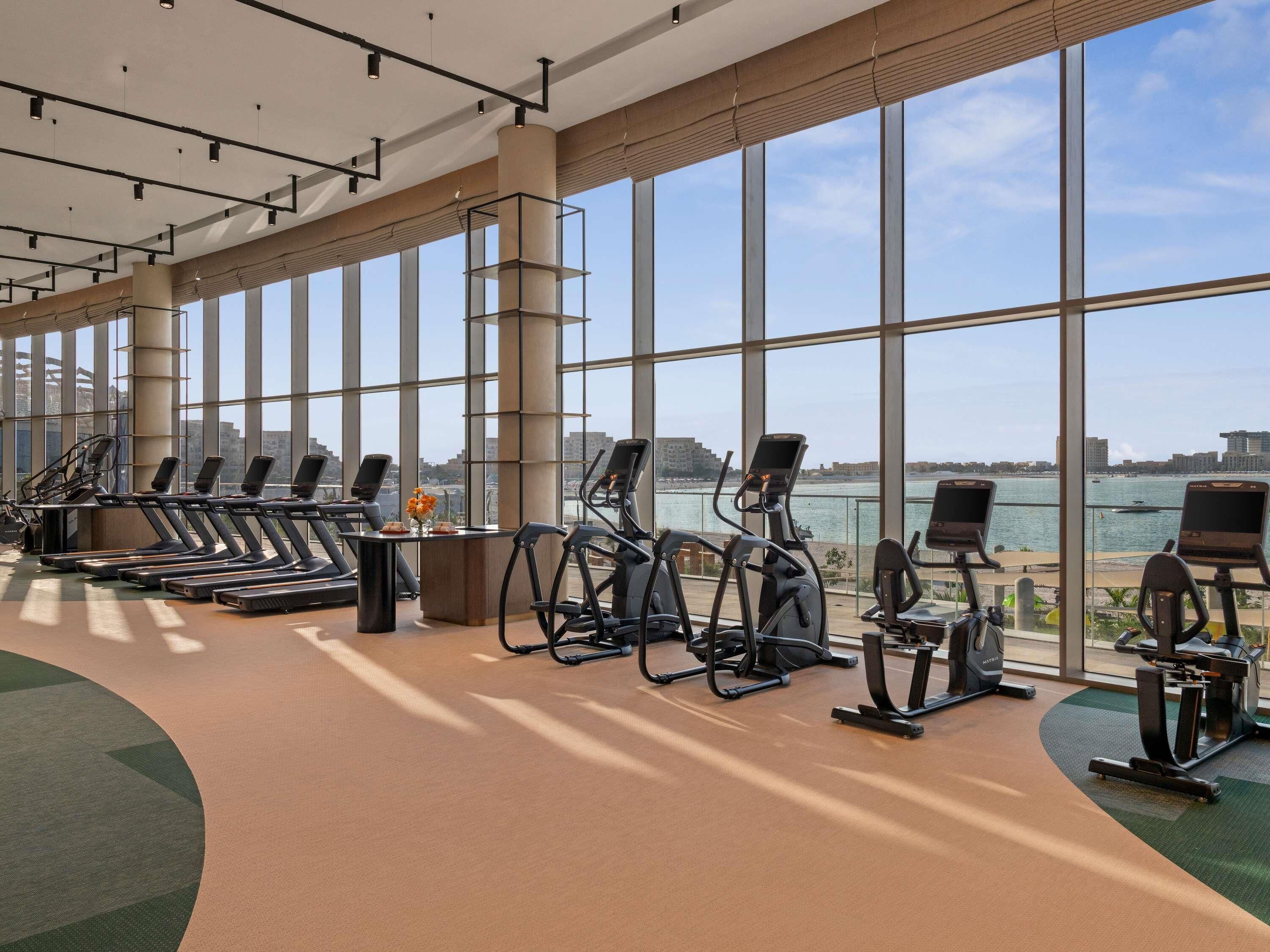 A gym with treadmills, elliptical machines, and stationary bikes, large windows, and a view of a body of water.