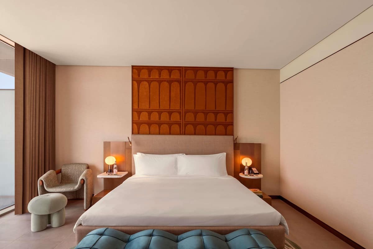 A hotel room with a large bed, two bedside tables with lamps, a chair, a small ottoman, and a decorative headboard.