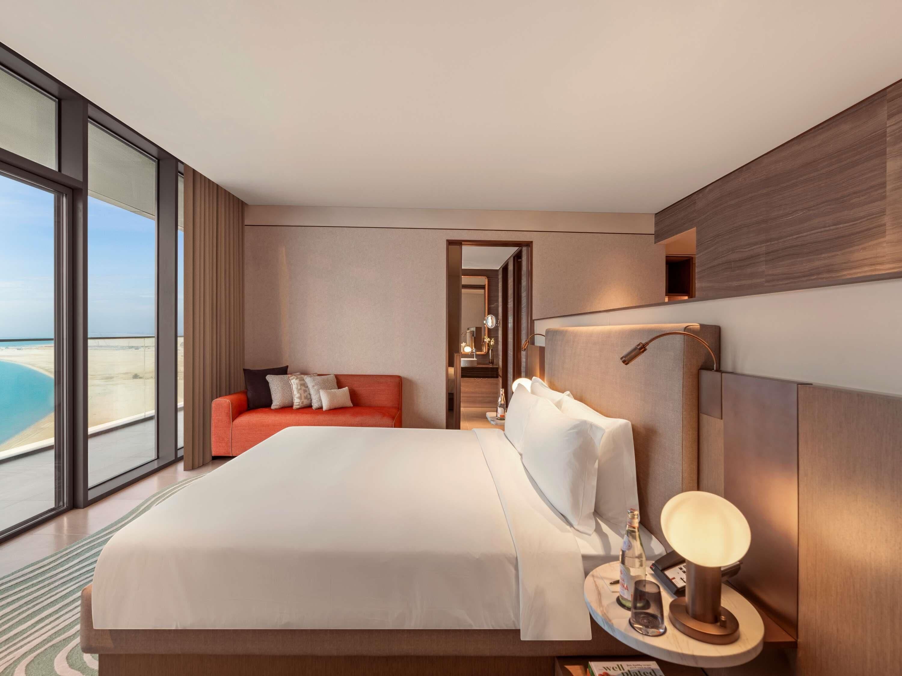 A modern hotel room with a large bed, a sofa, a bedside table with a lamp, and a view of the beach.