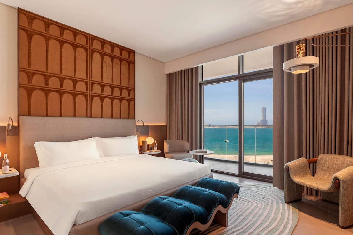 A hotel room with a large bed, a bench, a chair, a nightstand, a lamp, and a view of the sea.