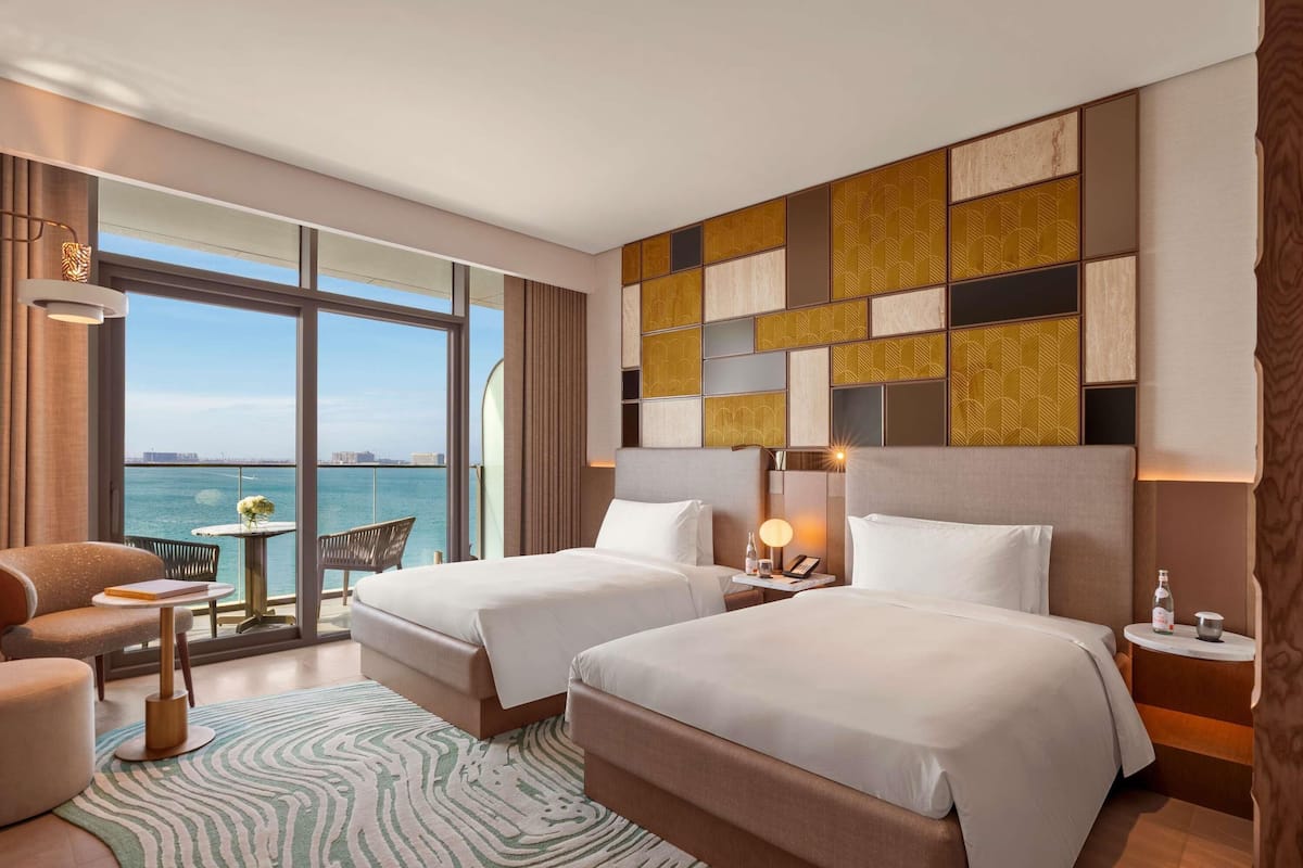A hotel room with two beds, a view of the sea, a table with a chair, and a patterned rug.