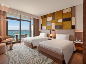 Habitación de hotel con dos camas, vista al mar, una mesa con una silla y una alfombra estampada.