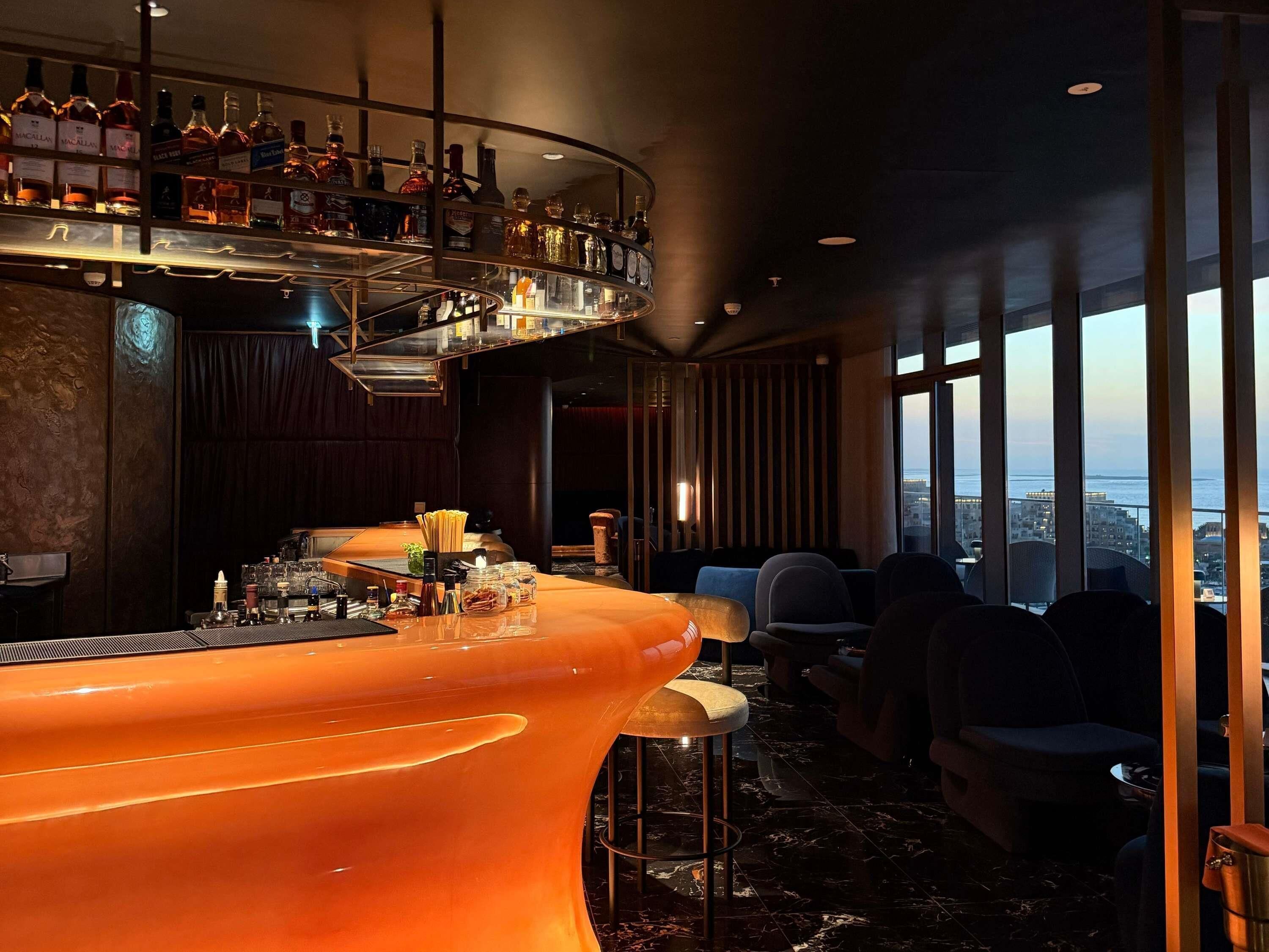 A bar area with a curved counter, shelves with bottles, and a view of the city through large windows.